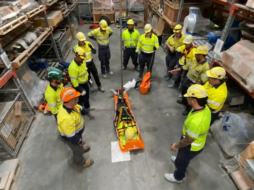 A training group gathered around a stretcher being lowered on a rope inside a warehouse
